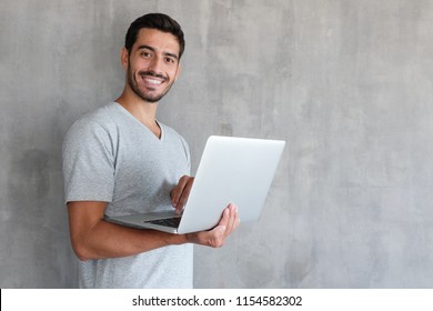 Indoor Portrait Of Young Man In T Shirt Standing Against Textured Wall With Copy Space For Ads, Holding Laptop And Looking At Camera With Happy Smile