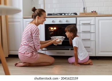 Indoor profile shot of smiling taking baking out of oven, her daughter standing near by and smelling delicious sweets, kid helping mother on kitchen, family posing at home while sitting on the floor.