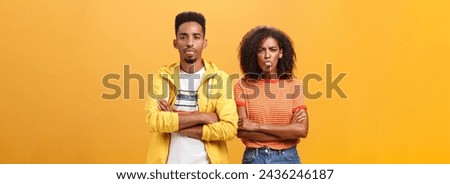 Indoor shot of african american siblings being displeased and annoyed showing bad tempber behaving childish sticking out tongue standing together with crossed hands on chest over orange background