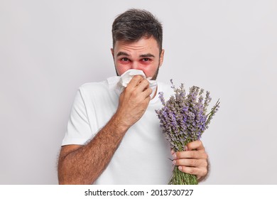 Indoor Shot Of Allergic Man Blows Nose In Handkerchief Has Sneezing And Rhinitis Allergy To Lavender Red Swelling Eyes Suffers From Unpleasant Symptoms Isolated On White Wall. Sickness Concept