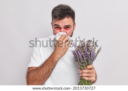 Indoor shot of allergic man blows nose in handkerchief has sneezing and rhinitis allergy to lavender red swelling eyes suffers from unpleasant symptoms isolated on white wall. Sickness concept
