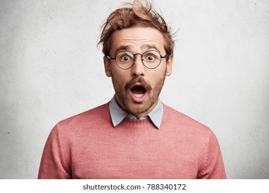 Indoor shot of amazed shocked young man with mustache, stares through round spectacles, being surprised to hear unexcpected news, can`t believe eyes, isolated over white concrete background.