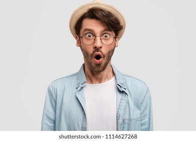 Indoor shot of amazed young Caucasian male hipster hears rumor from interlocutor, reacts on something, keeps mouth opened, feels excited, isolated on white background. Omg and surprisement concept