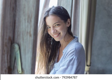 Indoor shot of amazing sophisticated young mixed raced woman wearing blue oversized shirt posing by window at light modern bedroom. Authentic lady looking at camera with cheerful pretty toothy smile.