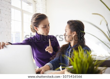 Indoor shot of angry young mother making warning sign, raising index finger, forbidding her daughter to spend so much time online, browsing internet instead of doing homework. Technology and addiction