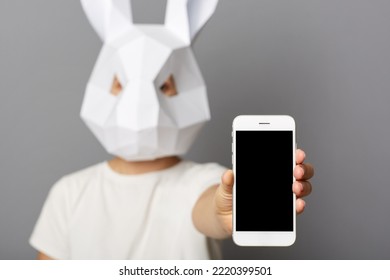 Indoor Shot Of Anonymous Woman Wearing White Shirt And Rabbit Mask Posing Isolated Over Gray Background, Showing Cell Phone With Empty Display With Copy Space For Advertisement.