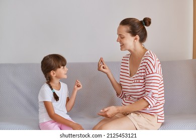 Indoor Shot Of Attractive Young Adult Woman Wearing Striped Shirt And Shorts Sitting On Sofa With Little Girl And Having Session Of Speech Therapy, Female Demonstrating Correct Sounds Pronunciation.