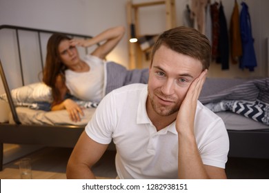 Indoor Shot Of Attractive Young Casually Dressed With Stubble Sitting On Floor In Bedroom, Looking And Smiling At Camera, His Wife Lazing In Bed, Not Ready To Get Up. Selective Focus On Man's Face