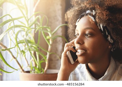 Indoor shot of attractive young dark-skinned woman, using cell phone for ordering pizza for dinner. Serious female having phone talk using electronic device, relaxing at home, looking through window - Powered by Shutterstock