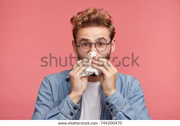 Indoor shot of bearded man has running nose, rubs\
nose with handkerchief, being ill, caught cold after long walk\
outdoors on rainy weather. Unhealthy allergic male sneezes isolated\
over pink wall