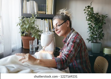 Indoor shot of beautiful elderly woman tailor sewing winter coat using stitching machine at her workplace, looking and smiling joyfully at camera, enjoying her hobby. People, occupation and job