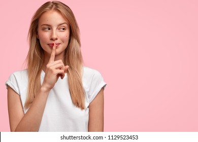 Indoor shot of beautiful female looks mysteriously aside, has intriguing look, asks to be quiet, dressed in casual clothes, stands against pink background with blank copy space for your advertisement - Shutterstock ID 1129523453