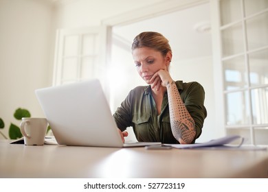 Indoor shot of beautiful female sitting at table and using laptop computer. Woman working seriously on laptop at home.