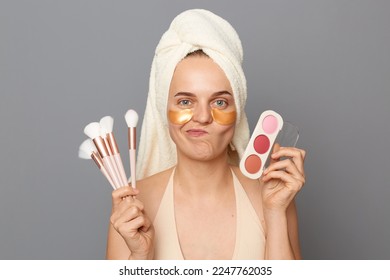 Indoor shot of beautiful woman holding shadows for eyelids, standing isolated over gray background, doing makeup, having confused expression, has no color on her palette. - Shutterstock ID 2247762035