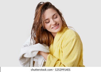 Indoor Shot Of Beautiful Woman Wipes Wet Hair With Towel, Dressed In Yellow Bathrobe, Stands Against White Background, Has Pleased Facial Expression, Has Shower At Home, Satisfied With New Shampoo