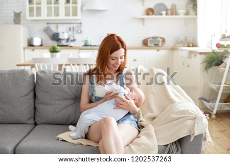 Indoor shot of beuatiful red haired European mom nursing toddler on comfortable couch with kitchen in background, looking at baby with love while feeding him formula from plastic bottle with teat