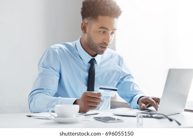 Indoor Shot Of Black Male Businessman Wearing Blue Shirt With Tie Using Laptop Computer In Office Holding Debit Card In His Hands, Entering Its Number While Filling In Online Form On Some Web-site