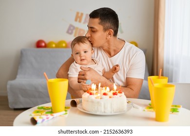 Indoor shot of brunette female man with infant baby celebrating first birthday, sitting at table with birthday cake and drink, father kissing his infant daughter, celebrating together.