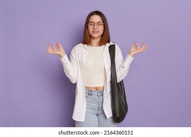 Indoor Shot Of Calm Relaxed Attractive Woman With Black Bag Wearing White Shirt And Jeans Posing Isolated Over Purple Background, Standing With Closed Eyes, Practicing Yoga To Calm Down.