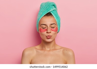 Indoor Shot Of Calm Relaxed Woman Has Fresh Healthy Skin, Keeps Eyes Shut, Wears Collagen Patches Under Eyes, Wears Turquoise Towel On Head, Keeps Lips Rounded, Gets Facial Treatment In Spa Salon