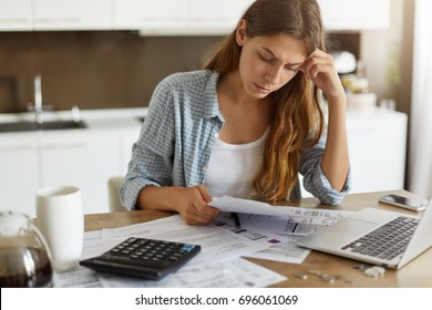 Indoor shot of casually dressed young woman holding papers in her hands, calculating family budget, trying to save some money to buy new bicycle to her son, having stressed and concentrated look - Shutterstock ID 696061069