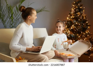 Indoor Shot Of Caucasian Woman Sitting On Cough With Her Daughter And Working On Laptop, Talking With Her Daughter Who Eating Pizza, Family Spending New Year Eve.