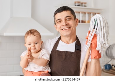 Indoor Shot Of Caucasian Young Adult Brunette Man Holding Infant Baby And Floor Mops For Cleaning Kitchen, Wearing Brown Apron And White T Shirt, Looking At Camera With Happy Facial Expression.