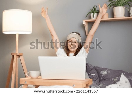 Indoor shot of charming optimistic happy woman wearing white t-shirt and blindfold sitting in bed in front of laptop, stretching her arms, waking up in high spirit.