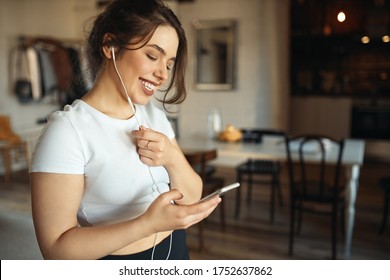 Indoor shot of charming young plus size woman in white top holding smart phone using headset while talking to friend via online video chat, smiling, making plans for weekend. People and technology - Powered by Shutterstock