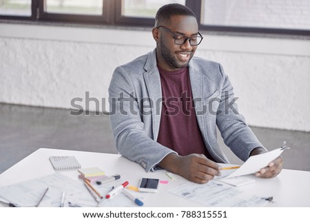Indoor shot of cheerful African American male enterpreneur sits at table surrounded with papers, studies financial report, rejoices raising sales and acheivement goals. Black businessman at work