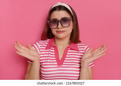 Indoor shot of confused woman wearing striped T-shirt and sunglasses posing isolated over pink background, being not sure, don't know the answer and the solution.