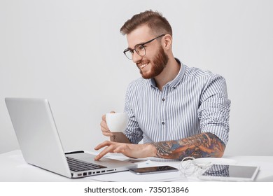 Indoor Shot Of Creative Business Worker With Tattooed Arms, Has Thick Beard, Holds Mug Of Tea, Looks Pleased Into Screen Of Laptop, Messages With Friends As Has Coffee Break. People, Modern Technology