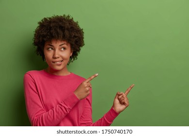 Indoor shot of curious African American woman bites lips, sees something good, indicates fingers at blank space on green background, shows place for your product, company banner or promotion - Shutterstock ID 1750740170