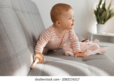 Indoor Shot Of Curious Charming Infant Baby Girl Wearing Striped Sleeper Posing On Gray Sofa At Home Alone, Looking Away, Finding Her Mother, Crawling.