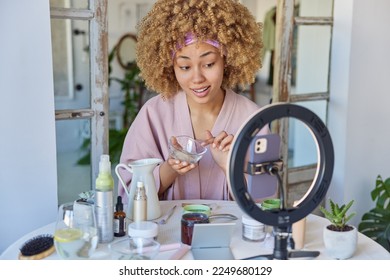 Indoor shot of curly haired adult woman dressed in bathrobe records video blog tells about cosmetic products for your beauty poses against blurred home interior. Blogging and cosmetology concept - Powered by Shutterstock