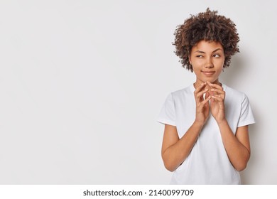 Indoor shot of curly haired young woman steepls fingers looks away has mysterious expression evil plan dressed casually isolated over white background copy space for your promotional content