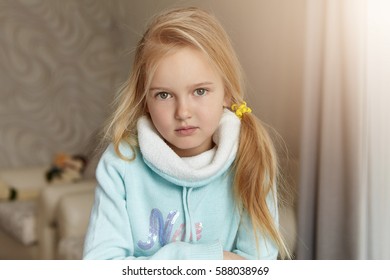 Indoor Shot Of Cute Blonde Little Girl Of European Appearance Wearing Her Hair In Messy Ponytail Posing Indoors Against Cozy Domestic Interior Background, Looking At Camera With Subtle Smile