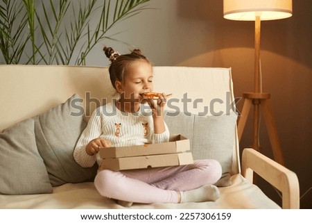 Indoor shot of cute funny child biting big slice pizza, sitting on sofa with crossed legs, enjoying tasty junk food, positive female kid with two bun hairstyle having dinner, eating fast food.