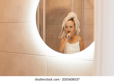 Indoor Shot Of Cute Girl Kid Brushing Teeth In Bathroom, Standing In Front Of The Mirror, Wrapped Her Head In Towel, Hygienic Procedures In The Morning Or Before Going To Bed