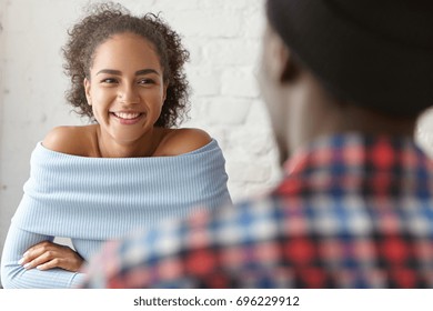 Indoor shot of cute mixed-race young couple having date, talking, enjoying nice time during lunch at cafe: pretty girl in blue top with open shoulders looking and smiling at unrecognizable man