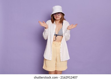 Indoor shot of dark haired European woman wearing swimsuit, shirt, shorts and panama, shrugging shoulders and dont know what to do, posing isolated over purple background.