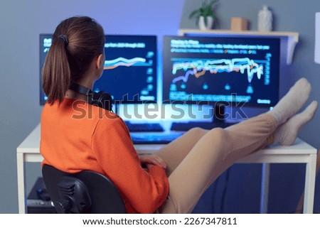 Indoor shot of dark haired female stock brokers wearing casual clothing is working in the home office by pc, sitting with headphones on neck and holding hands on table, looking at graphs on monitors.