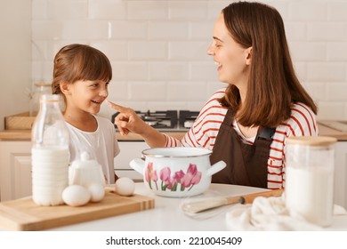 Indoor Shot Of Dark Haired Happy Satisfied Woman Baking Cake Or Pastries With Cute Little Daughter In Kitchen, Doing Bakery In Modern Kitchen, Having Fun.