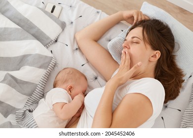 Indoor shot of of dark haired sleepy yawning tired female mother covering mouth, woman wearing white casual t shirt, breast feeding her infant, happy motherhood.