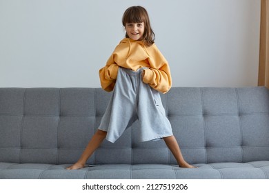 Indoor shot of dark haired smiling little girl wearing oversized yellow hoodie and gray short standing with legs wide apart, looking at camera, posing alone at home. - Shutterstock ID 2127519206