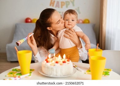 Indoor shot of dark haired woman with funny baby celebrating first birthday, sitting at table with birthday cake and drink, mother kissing her infant daughter, celebrating together.
