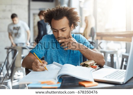Indoor shot of dark-skinned male student with African hairstyle wearing blue casual T-shirt sitting at university canteen eatting hamburger reading attentively book underline necessary information