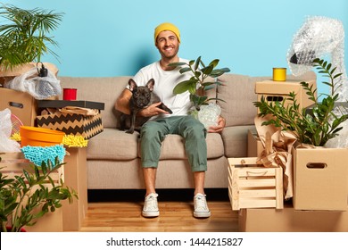 Indoor shot of delighted carefree house owner sits on couch and hugs pet, rests in own apartment, being in high spirit, celebrates move, has fun at home much household stuff around. Relocation concept