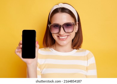Indoor shot of delighted dark haired young woman wearing striped t-shirt posing isolated over yellow background, showing mobile phone with empty display for advertisement.