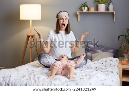 Indoor shot of despair woman wearing pajama and blindfold with her baby daughter in the bedroom sitting on bed, being tired to spend all hours with little child, screaming.
